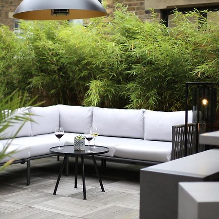 Middle Eight - Covent Garden - Preferred Hotels And Resorts London Exterior photo The photo shows an outdoor seating area featuring a modern gray sectional sofa with plush cushions. In front of the sofa is a small, round black table with two glasses of wine on it, accompanied by a decorative plant or vase. Surrounding the area are